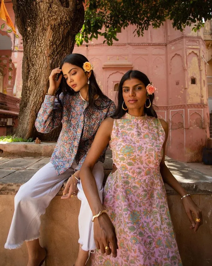 Two models in floral outfits beside a tree in front of a pink building